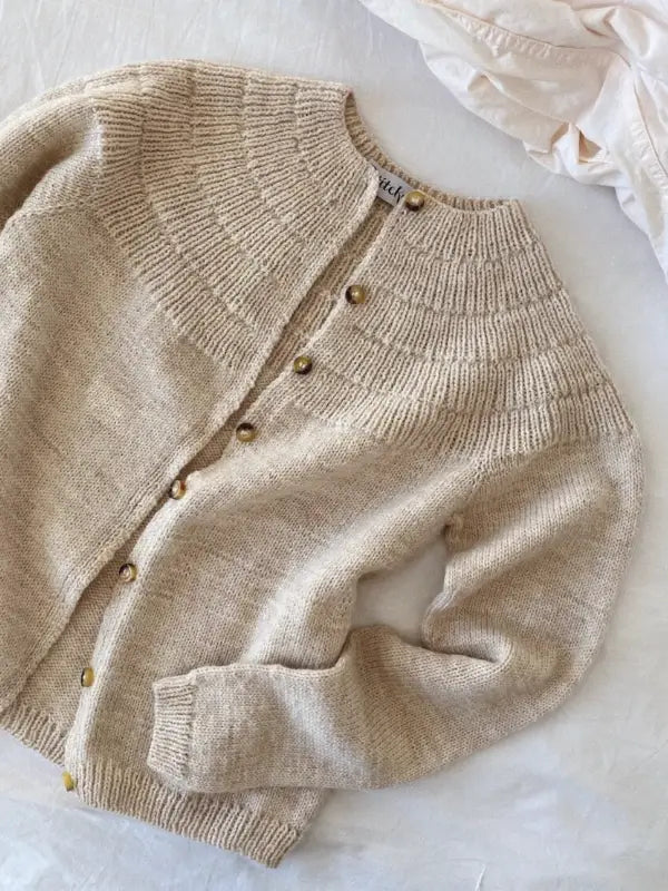 A beige cardigan inspired by PetiteKnit - Anker's Cardigan MY SIZE neuleohje features a ribbed circular yoke and brown buttons. It's laid flat on a white surface next to cream-colored fabric.