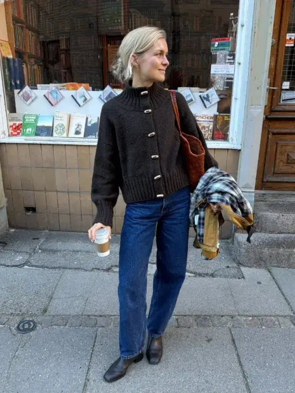 A person wears a dark brown PetiteKnit Hannah Jacket neuleohje -Suomi knit with a high collar and prominent buttons, paired with blue jeans and black boots.