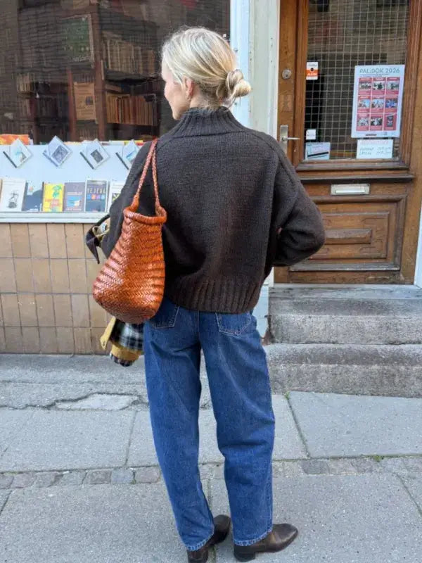 A person wearing a dark brown PetiteKnit Hannah Jacket neuleohje -Suomi knitted sweater with a high collar and blue jeans, carrying a woven brown shoulder bag.