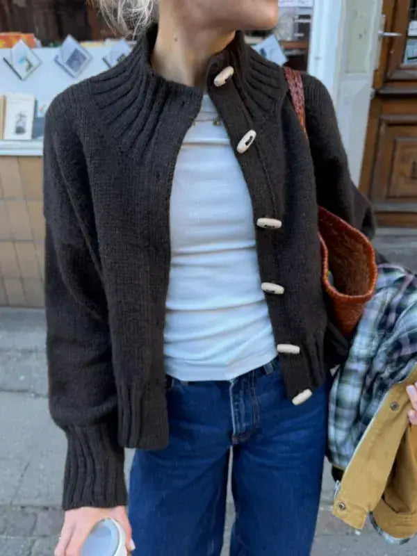 A person wearing a dark brown PetiteKnit Hannah Jacket neuleohje -Suomi knitted cardigan with a ribbed high collar and light-colored toggle buttons over a white t-shirt and blue jeans.