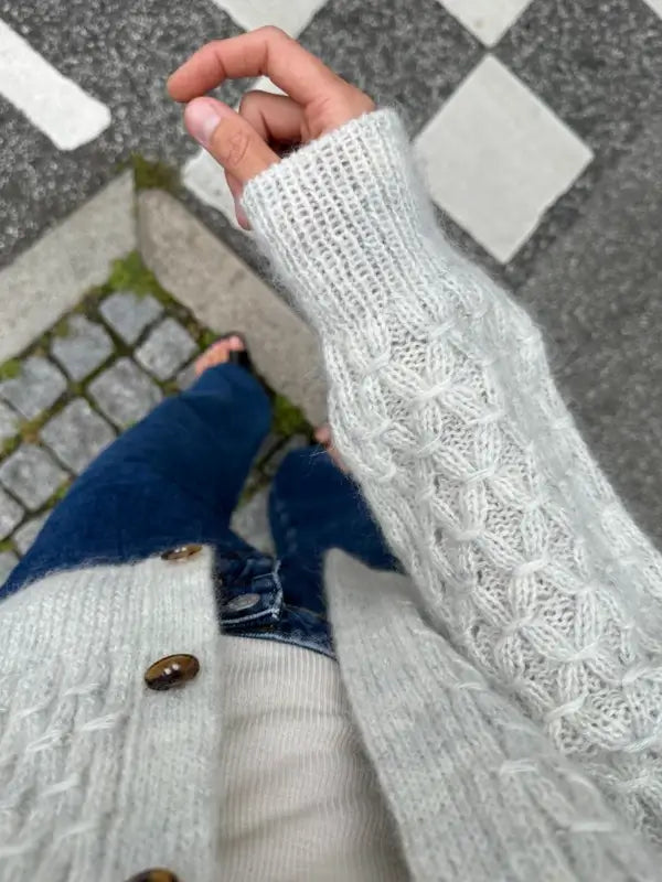 Wearing the PetiteKnit Jenny Jacket V-neck sweater, a person stands on stone pavement near a crosswalk. The photo, taken from above, highlights the textured knit details of the light gray neuleohje.