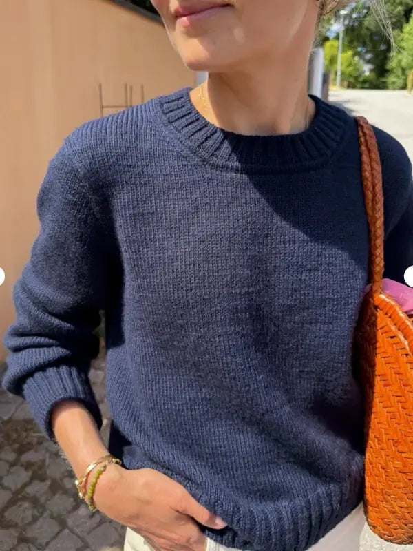 A person wearing a navy blue PetiteKnit Vega Sweater neuleohje and light pants stands outside, holding a woven brown bag. Sunlight highlights their face and sweater, perfectly showcasing the classic design of the PetiteKnit Vega Sweater neuleohje.
