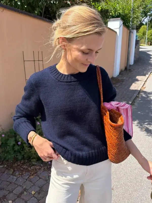 A woman wearing a navy PetiteKnit Vega Sweater neuleohje and white pants stands on a sunny sidewalk, holding a woven brown bag with a pink-striped item inside. She looks down and smiles slightly, with greenery and a peach wall behind her.