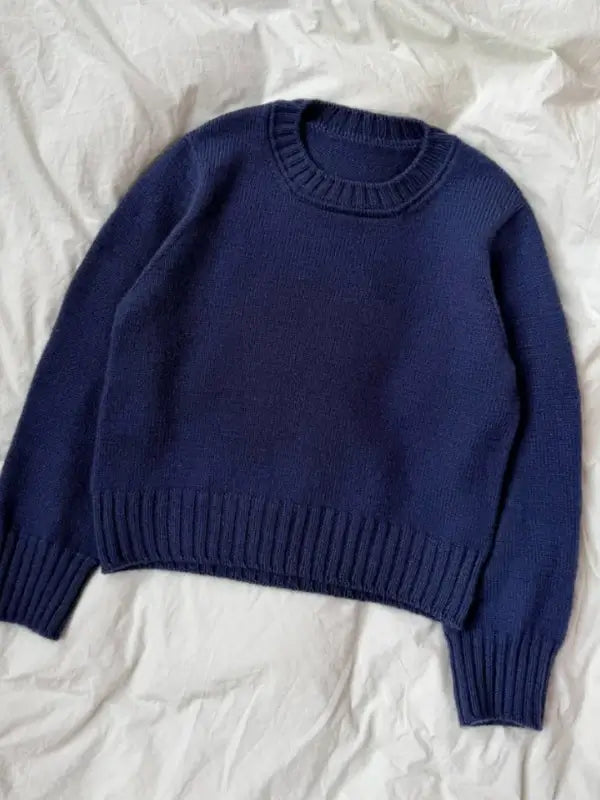 A plain, long-sleeved navy blue sweater, crafted with the PetiteKnit Vega Sweater neuleohje pattern, is laid flat on a wrinkled white fabric background.