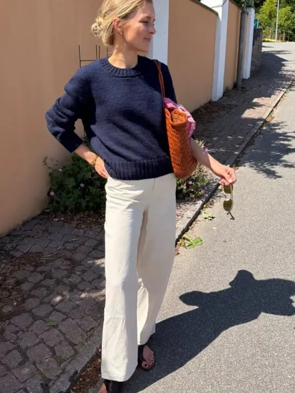 A woman stands on a sunny sidewalk wearing the PetiteKnit Vega Sweater neuleohje, its navy knit texture adding elegance to her look. She pairs it with cream wide-leg pants and sandals, carrying a woven tote bag and holding sunglasses, with her hair in a messy bun.