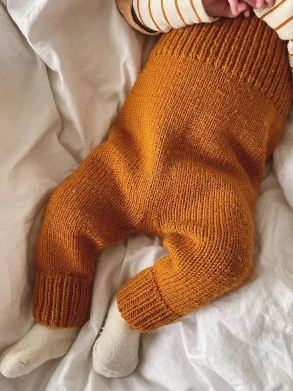 A baby wearing knit pants made from the PetiteKnit Willum’s Overalls neuleohje - Suomi pattern in a mustard hue, paired with white socks, lies on a white blanket. Only the baby’s legs, feet, and part of their torso are visible.