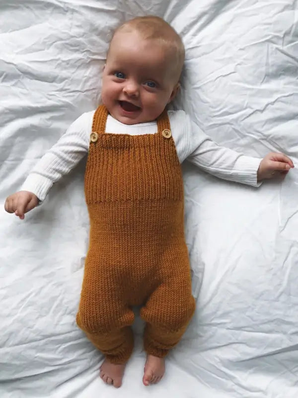 A smiling baby in a white long-sleeve shirt and mustard-colored knitted overalls made from the PetiteKnit Willum’s Overalls neuleohje - Suomi lies on a white blanket, looking up at the camera.