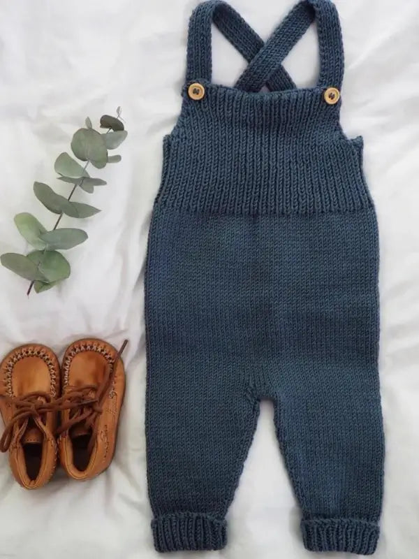 PetiteKnit Willum’s Overalls neuleohje - Suomi in navy blue, paired with brown baby moccasins and a sprig of eucalyptus, are arranged on a white fabric background. This charming setting showcases the cozy design of the PetiteKnit Willum’s Overalls, perfect for little ones.