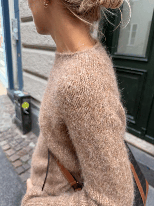 A person with light hair in a loose bun wears the Petiteknit IVY SWEATER neuleohje and carries a brown-strap bag while walking on a city street lined with stone pavement and classic buildings.