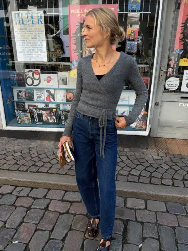 A woman wearing a gray PetiteKnit Lucy Wrap sweater, blue jeans, and sandals stands on a cobblestone street, holding a book and wallet. Behind her is a charming shop adorned with posters and records in the window, completing this scene inspired by the PetiteKnit Lucy Wrap - English neuleohje.