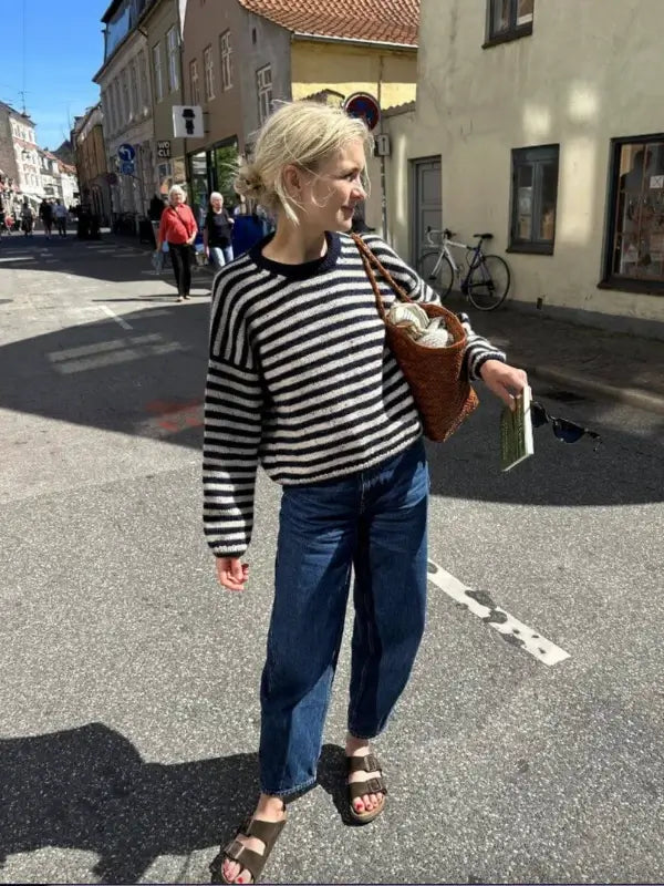 Wearing a black and white striped PetiteKnit Olga sweater neuleoohje with blue jeans and sandals, a person stands on a sunny street holding a brown bag and a book while looking to the side.