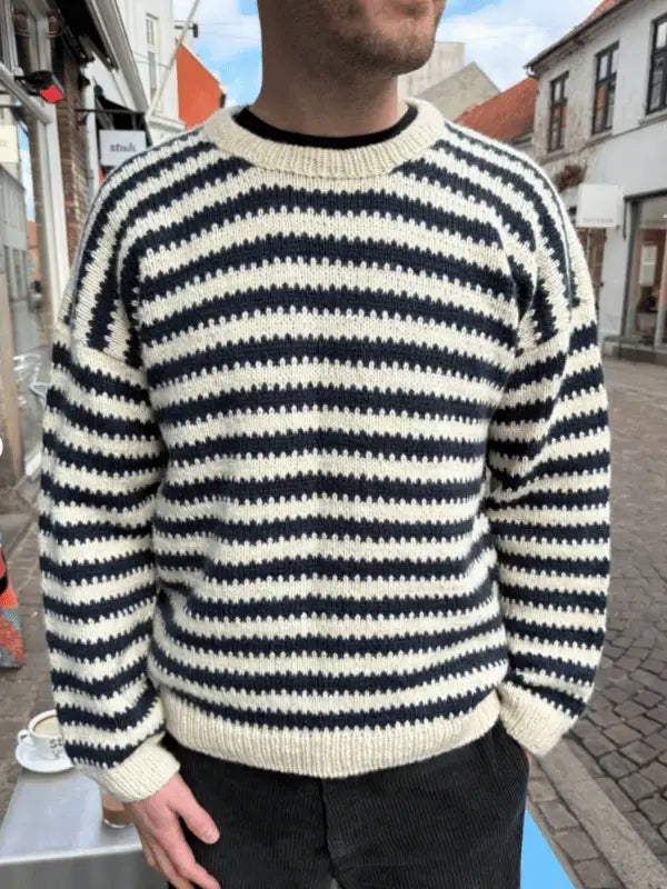 A person wearing a black and white PetiteKnit Otto Sweater neuleohje stands on a cobblestone street with buildings behind them. The ylhäältä alas neulottava paita has classic stripes, and they stand with hands at their sides, head slightly turned.