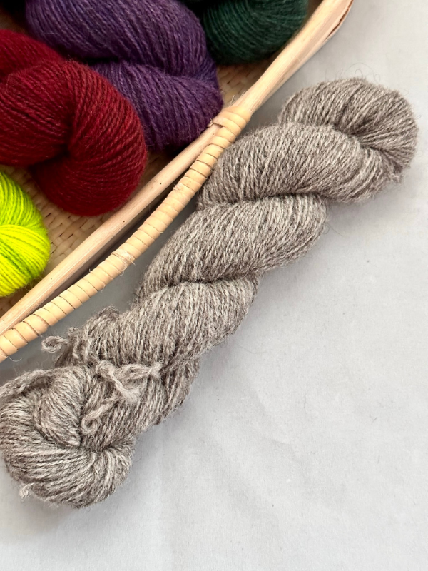 A skein of light gray WoolWomen Huiku yarn rests on a light surface beside a woven basket filled with red, purple, green, and yellow skeins.