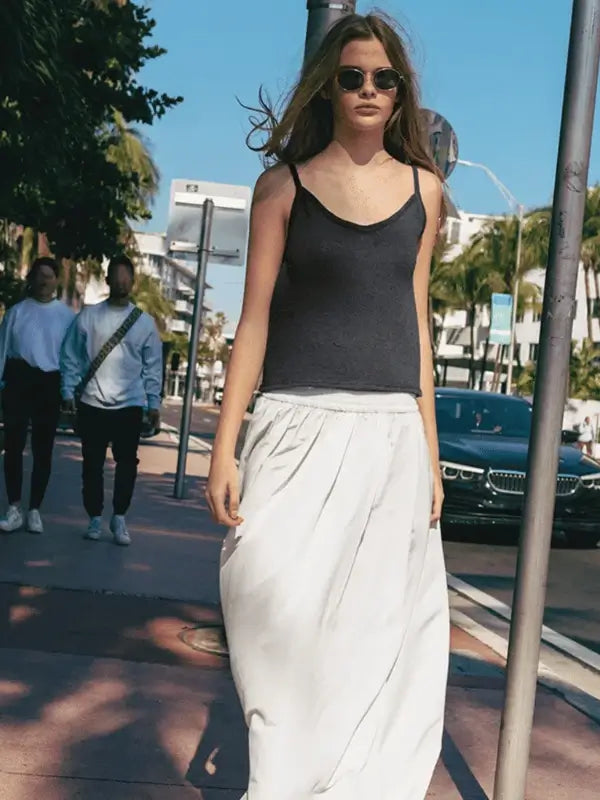 A woman wearing the Sandnes Garn Clementine Top (pattern in English, PDF) made with Sandnes Garn Tynn Line walks down a sunny city street, knitting needles in hand and people behind her.