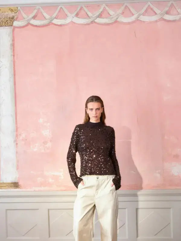 A person wearing a long-sleeved dark Sandnes Garn Isla Sweater – neulesetti Paljett covered in sequins with light-colored trousers.