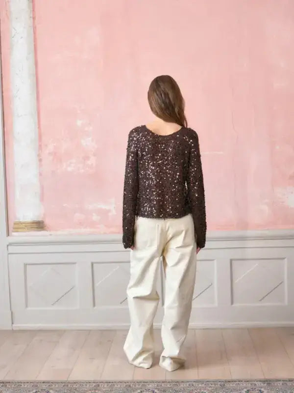A person seen from behind wearing a dark brown Sandnes Garn Isla Sweater – neulesetti Paljett with long sequined sleeves and loose-fitting cream trousers.