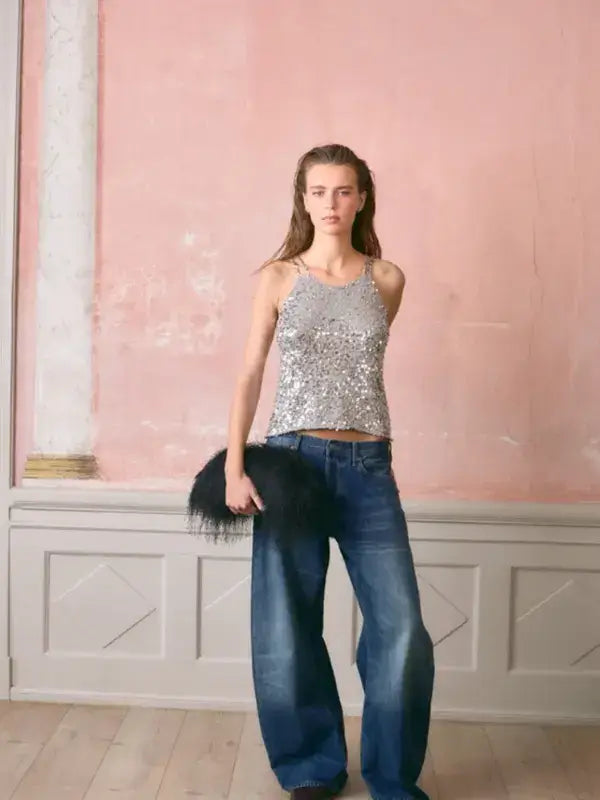 A model wearing a silver sequined Sandnes Garn Nia top – paljettitopin neulesetti and wide-leg blue jeans, holding a black feathered clutch.