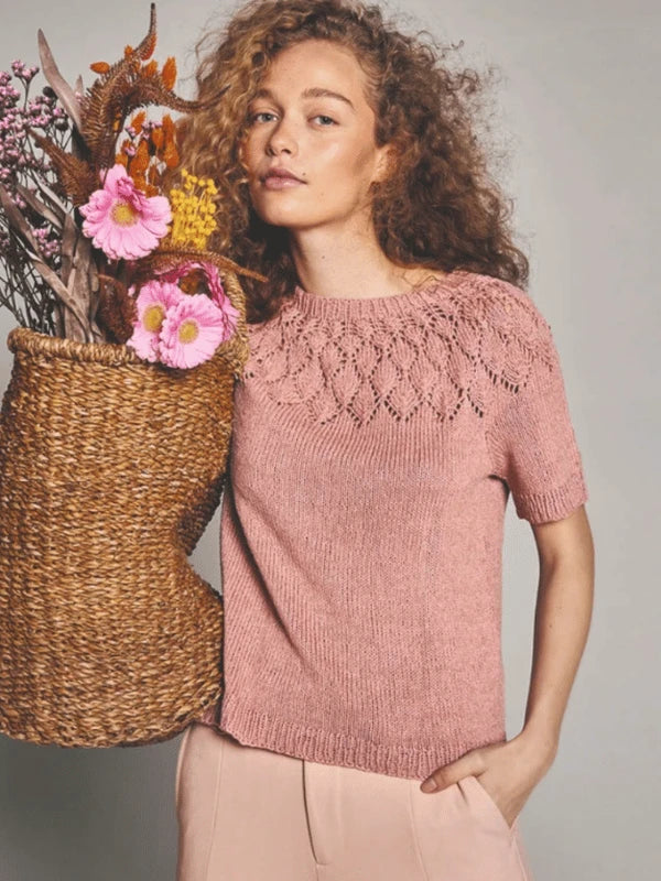 A woman with curly hair wears a pink, short-sleeved Sandnes Garn pitsineulepaita knit top and holds a large basket of pink, yellow, and brown flowers while standing against a plain background.