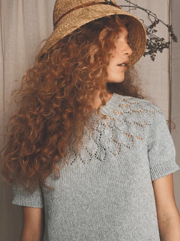 A person with long, curly red hair wears a straw hat and a light gray Sandnes Garn pitsineulepaita with a lacy leaf-patterned neckline, looking to the side against a neutral background.