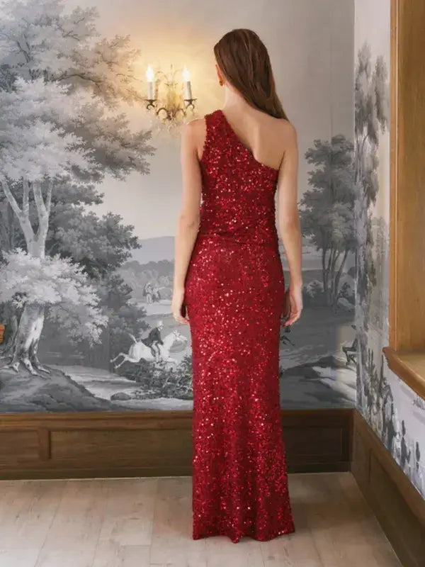 A woman stands with her back to the camera wearing a floor-length, one-shoulder red sequined Sandnes Garn Vale Top – neulesetti Paljett dress.