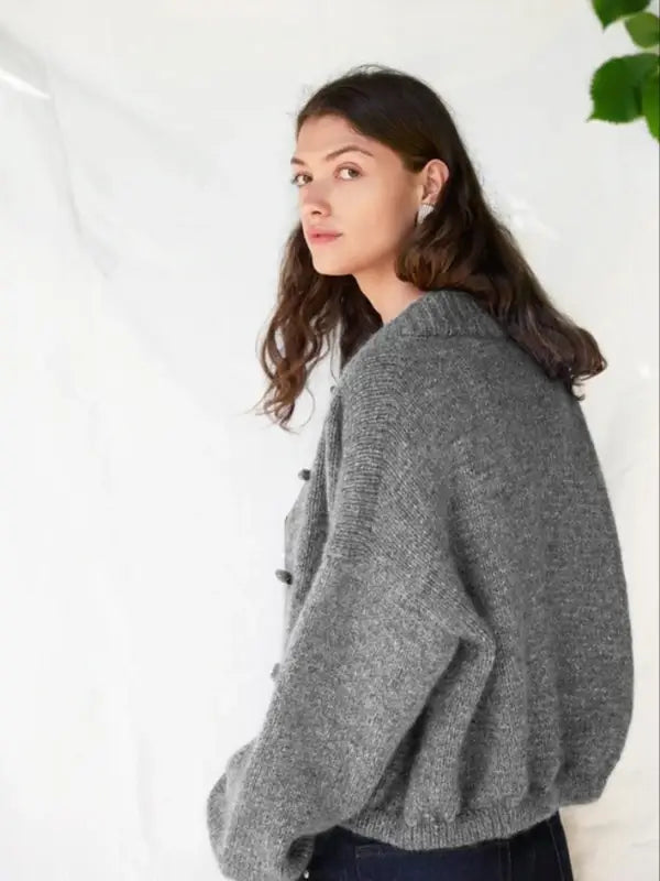 A young woman with long brown hair wears a loose grey DIY Bomber Jacket, crafted from Sandnes Garn 2606-08 (neuleohje Suomi, PDF), styled with dark pants and standing sideways, looking at the camera against a plain white background.