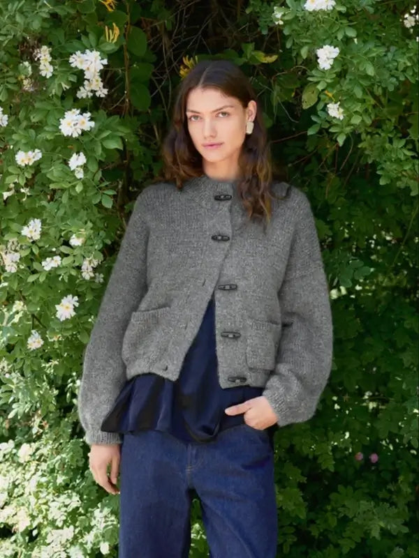 Wearing the Sandnes Garn 2606-08 DIY Bomber Jacket – neuleohje Suomi (PDF), a person with long brown hair stands in front of leafy green bushes with white flowers. The stylish gray, buttoned jacket is layered over a dark blouse and blue jeans, creating a chic look.