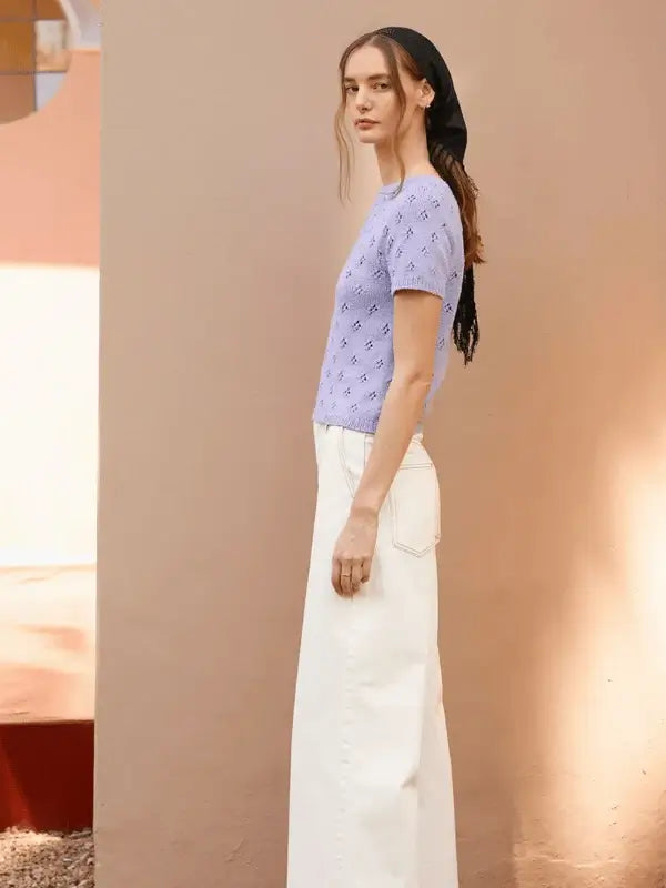 A woman stands in profile against a beige wall wearing the Elodie Tee neuleohje ENG, knitted with Sandnes Garn Line in light purple, paired with high-waisted white wide-leg pants and a black headscarf.