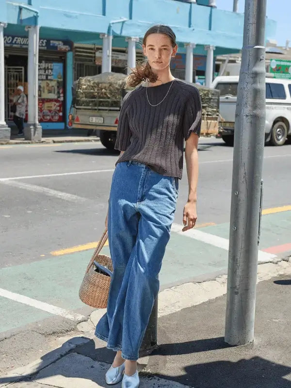 A woman stands on a city sidewalk wearing the Elva tee neuleohje ENG, knitted with Sandnes Garn Line, high-waisted wide-leg jeans, and white shoes. She carries a woven bag with her hair tied back; shops and vehicles are in the background.