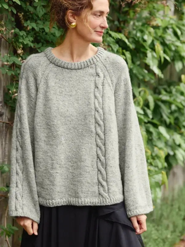 A person wearing a loose, light gray sweater made from the Sandnes Garn 2606-10 DIY Laura Sweater pattern—available as a Finnish-language PDF knitting guide—paired with a black skirt stands outdoors near a leafy green fence, looking to the side and smiling.