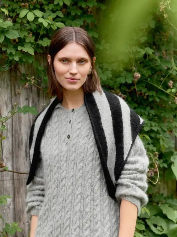 A woman with brown hair in a gray cable-knit sweater stands outside, smiling softly, with the Sandnes Garn 2606-06 DIY Mango Scarf–neuleohje suomeksi draped over her shoulders. Green leaves and a wooden fence can be seen in the background.