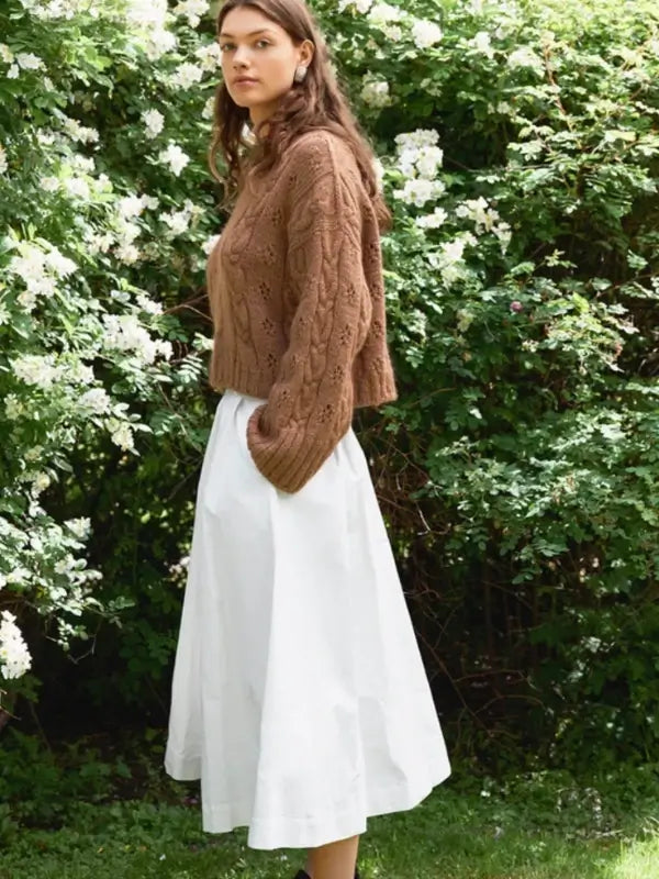 A woman stands outdoors near bushes with white flowers, wearing a cozy brown sweater made using the Sandnes Garn 2606-11 Junia Sweater – neuleohje suomeksi PDF knitting pattern, paired with a white midi skirt. She has long, wavy brown hair and gazes calmly toward the camera.
