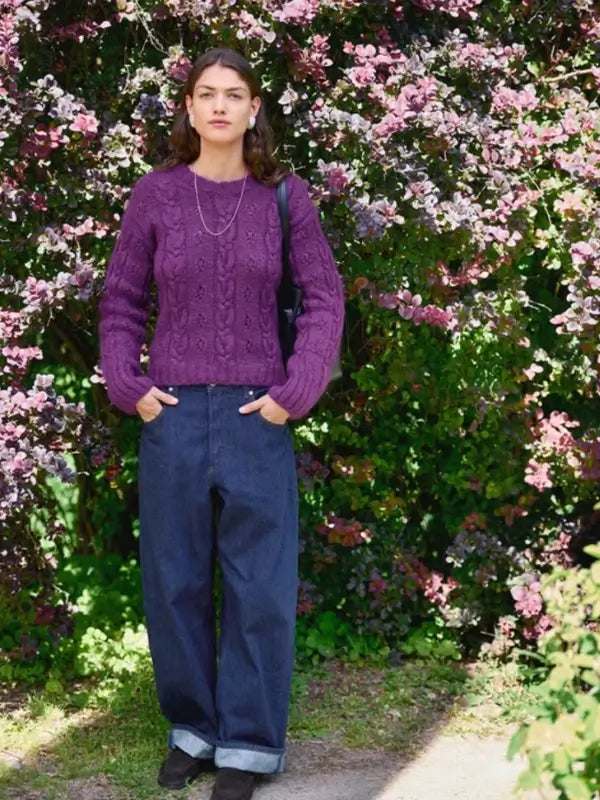 A woman stands outdoors in front of blooming pink bushes, wearing a sweater made from the Sandnes Garn 2606-11 Junia Sweater – neuleohje suomeksi PDF pattern, paired with loose blue jeans, black shoes, and a necklace, her hands casually tucked in her pockets.