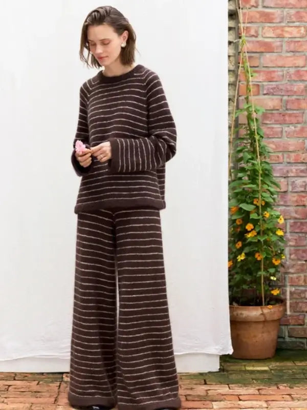 A person stands on a brick patio near a potted plant with orange flowers and a brick wall, wearing the cozy Sandnes Garn Riley Sweater & Riley Pants – neuleohje suomeksi, PDF set. The outfit features a loose, brown sweater and matching wide-leg pants with white horizontal stripes, making it perfect for anyone looking for Finnish knitting patterns in PDF format.