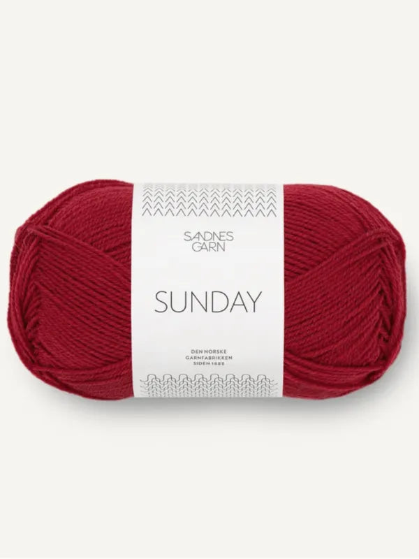 A skein of Sandnes Garn Sunday, fingering weight yarn in deep red, with a white label featuring minimalist black text and patterns, set against a plain, light background.