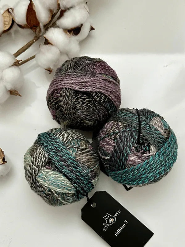 Three balls of Schoppel-Wolle Edition 3 superwash merino yarn in grey, green, and purple are on a white surface next to a cotton branch. One ball shows the product’s black label.