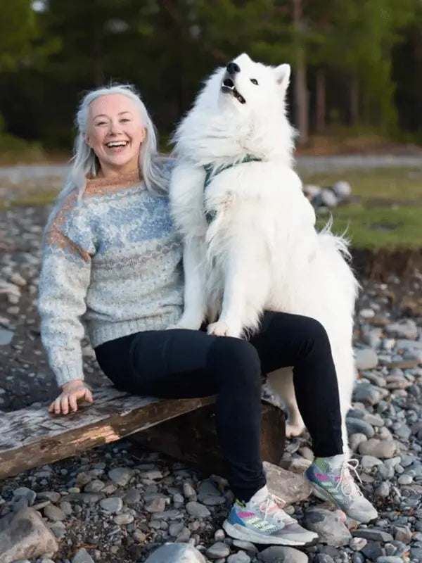 A smiling woman in a Sisu Knitwear Lohineule neuleohje Suomi PDF sweater sits on a wooden bench outdoors, with a large fluffy white dog resting its paws on her lap amid rocky ground and trees in the background.