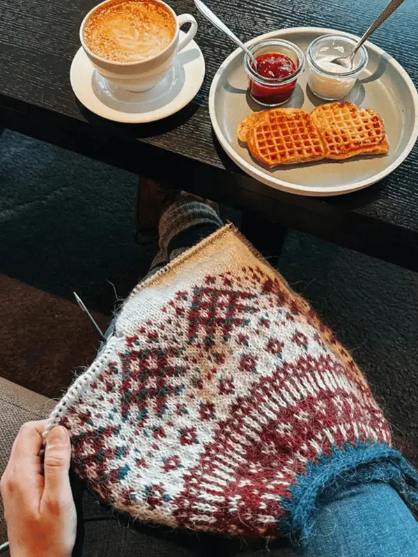 A person is knitting with the Sisu Knitwear Rovaneule neuleohje Suomi PDF in their lap. On a nearby table, there’s a cup of coffee and a plate holding two waffles topped with strawberry jam and cream.