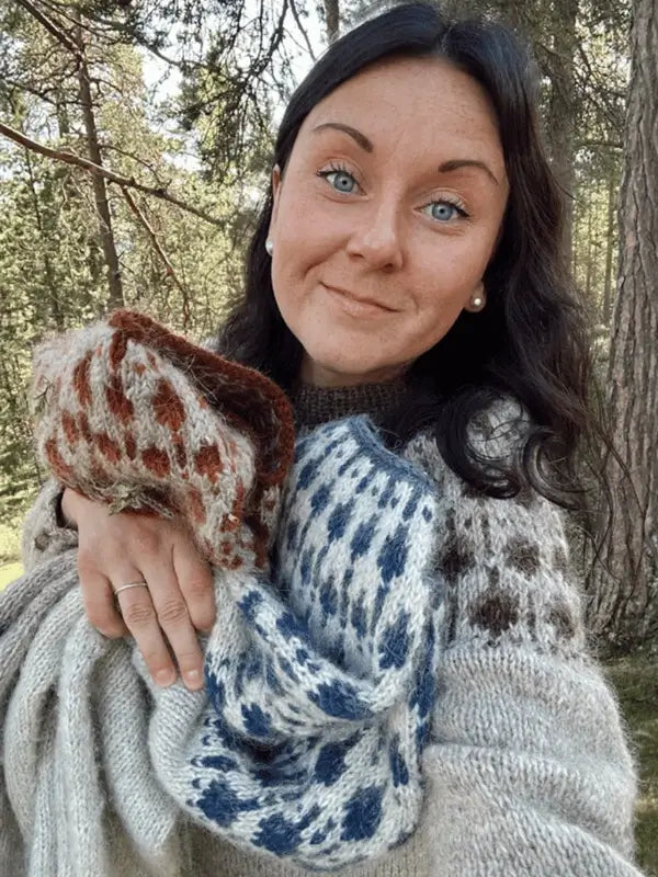 A woman with long dark hair stands in a forest, smiling and holding hand-knitted sweaters. She wears a cozy Sisu Knitwear Terhoneule neuleohje Suomi PDF-inspired sweater with a pyöreä kaarroke and pearl earrings.