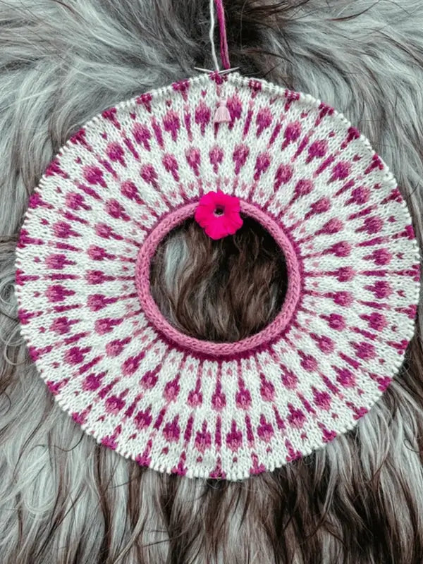 A circular knitted Sisu Knitwear Vadelmaneule neuleohje piece in pink and white dots hangs against a furry gray-brown background, featuring a small pink flower by the center. Design available as a Suomii PDF.