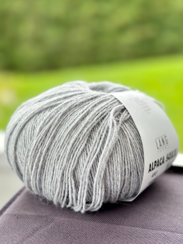 A ball of Lang Alpaca Soxx 4ply in light gray with a white label rests on a dark surface—perfect for your next Mittums lapasten ohje (PDF) project—with a blurred green background.