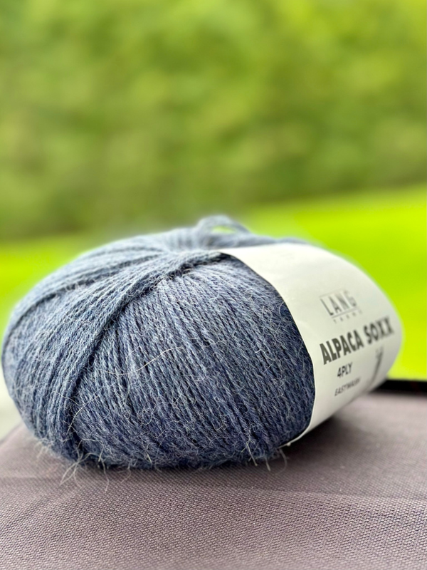 A skein of Lang Alpaca Soxx 4PLY lies on a dark surface beside the Mittums lapasten ohje (PDF), perfect for your next project, with a soft green outdoor blur in the background.