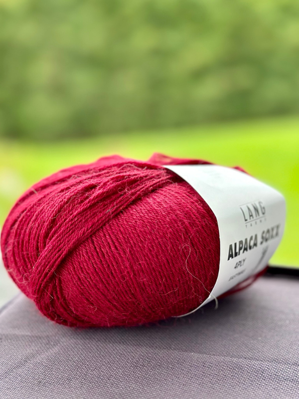 A close-up of Lang Alpaca Soxx 4ply in deep red with a white label on gray, set outdoors—perfect for cozy mittens with the Mittums lapasten ohje (PDF) knitting pattern.