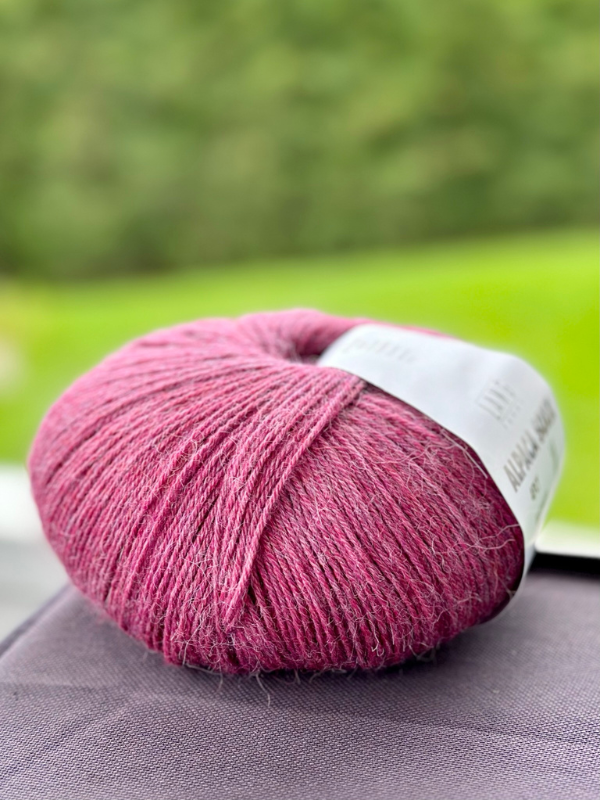 A close-up of Mittums lapasten ohje (PDF) + Lang Alpaca Soxx 4ply pink fingering yarn with a white label on a dark surface, against a blurred background of green grass and outdoor foliage.