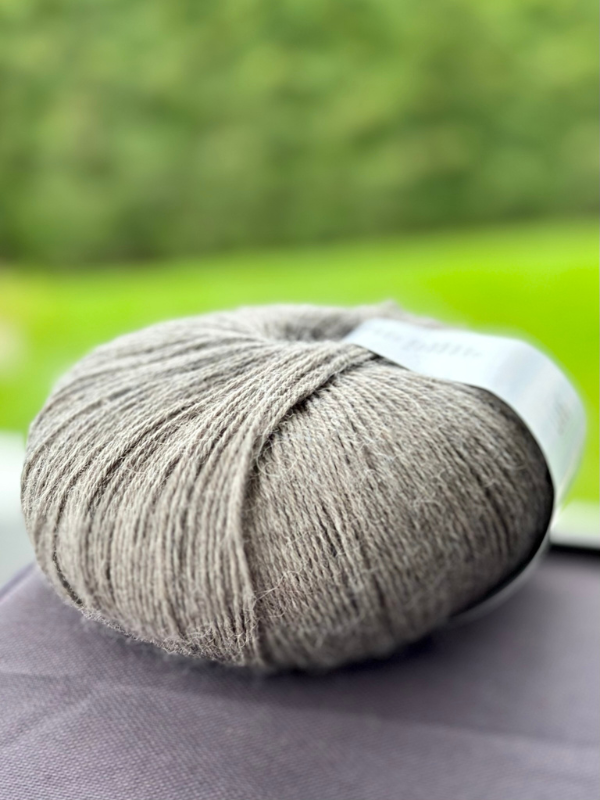 A close-up of Lang Alpaca Soxx 4ply yarn from the Mittums lapasten ohje (PDF) kit rests on a dark surface with a blurred green background.