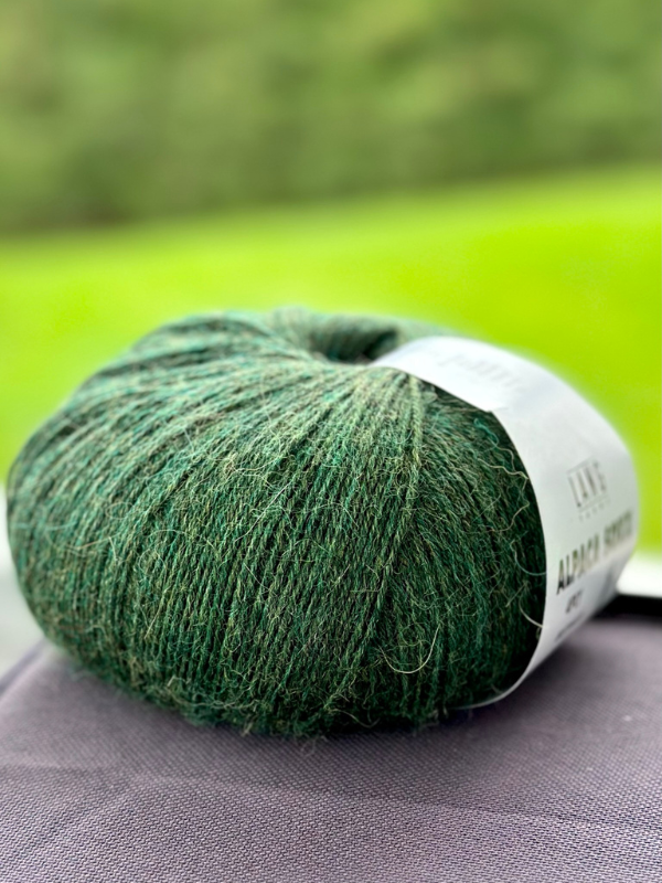 A ball of Lang Alpaca Soxx 4ply yarn from Mittums lapasten ohje (PDF) rests on a dark fabric surface, with a blurred green outdoor background.