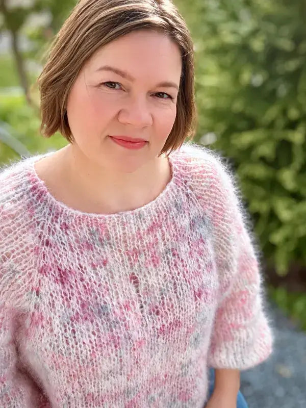 A woman with short brown hair, dressed in a soft and fluffy sweater made from the Hattara neuleohje pattern in light pink and white Fluff Mohair, is smiling gently outdoors with greenery in the background.