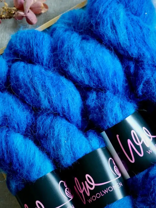 Three skeins of vibrant blue WoolWomen Alpaca Glimmer yarn with black labels reading Woolwoven in pink script, arranged closely together on a gray surface.