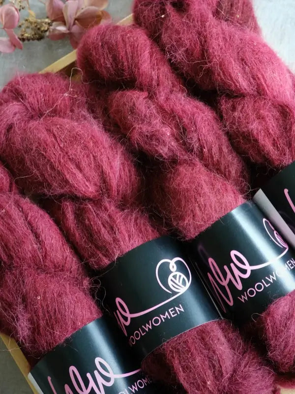 Deep red skeins of WoolWomen Alpaca Glimmer are neatly arranged, each wrapped with a black label that reads Woolwomen. Pink flowers are partly visible in the upper left corner of the image.
