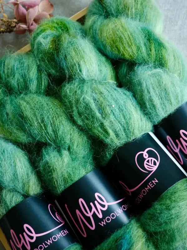 Five skeins of fluffy, green WoolWomen Alpaca Glimmer yarn are stacked together on a surface, with dried flowers in the corner. This käsinvärjätty alpakkalanka offers luxurious softness and vibrant color—perfect for your next special project.
