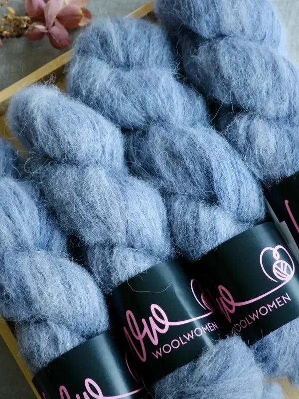Four skeins of soft, fluffy blue WoolWomen Alpaca Glimmer yarn are displayed side by side. Each skein features a black label with pink writing that reads WoolWomen. A small pink flower in the background adds a delicate touch to this luxurious käsinvärjätty alpakkalanka (hand-dyed alpaca yarn) collection.